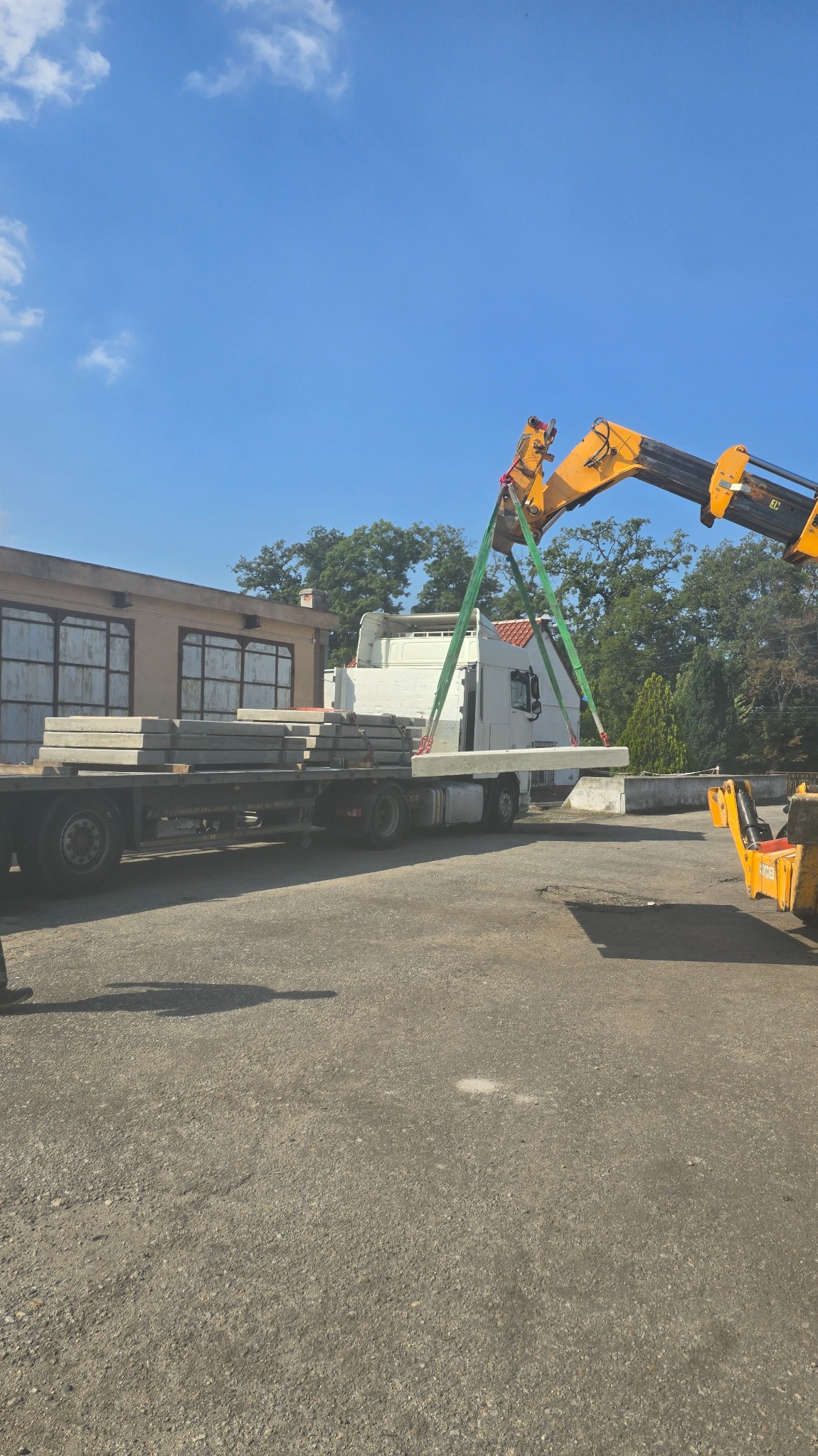Żuraw przenosi betonową płytę drogową z ciężarówki na plac. Widoczne budynki w tle i błękitne niebo. Transport i rozładunek płyt.