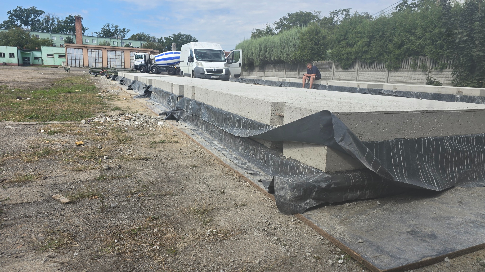 Ułożone płyty betonowe na placu budowy, zabezpieczone folią. W tle betoniarka i budynek. Mężczyzna siedzi na płycie, używając telefonu. Dzień, pochmurne niebo.