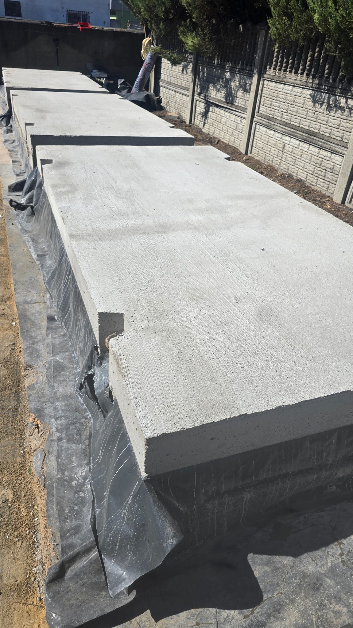Ułożone płyty drogowe z betonu na czarnej folii, widoczne charakterystyczne wycięcia na krawędziach, w tle betonowe ogrodzenie i zieleń.