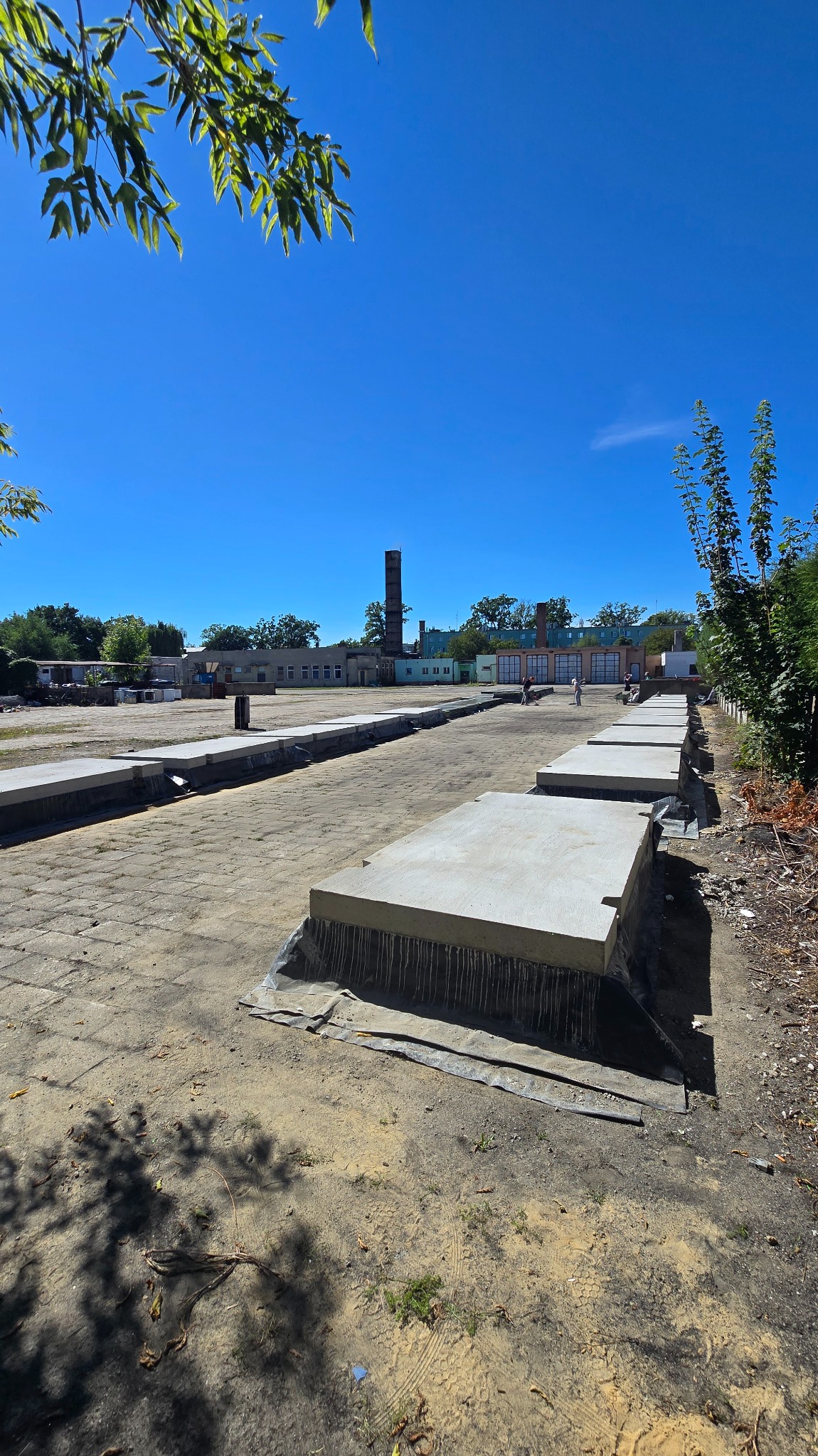 Układanie betonowych płyt drogowych na placu budowy w słoneczny dzień. Widoczny fragment placu z ułożonymi płytami, budynek w tle i komin. Prace w toku.