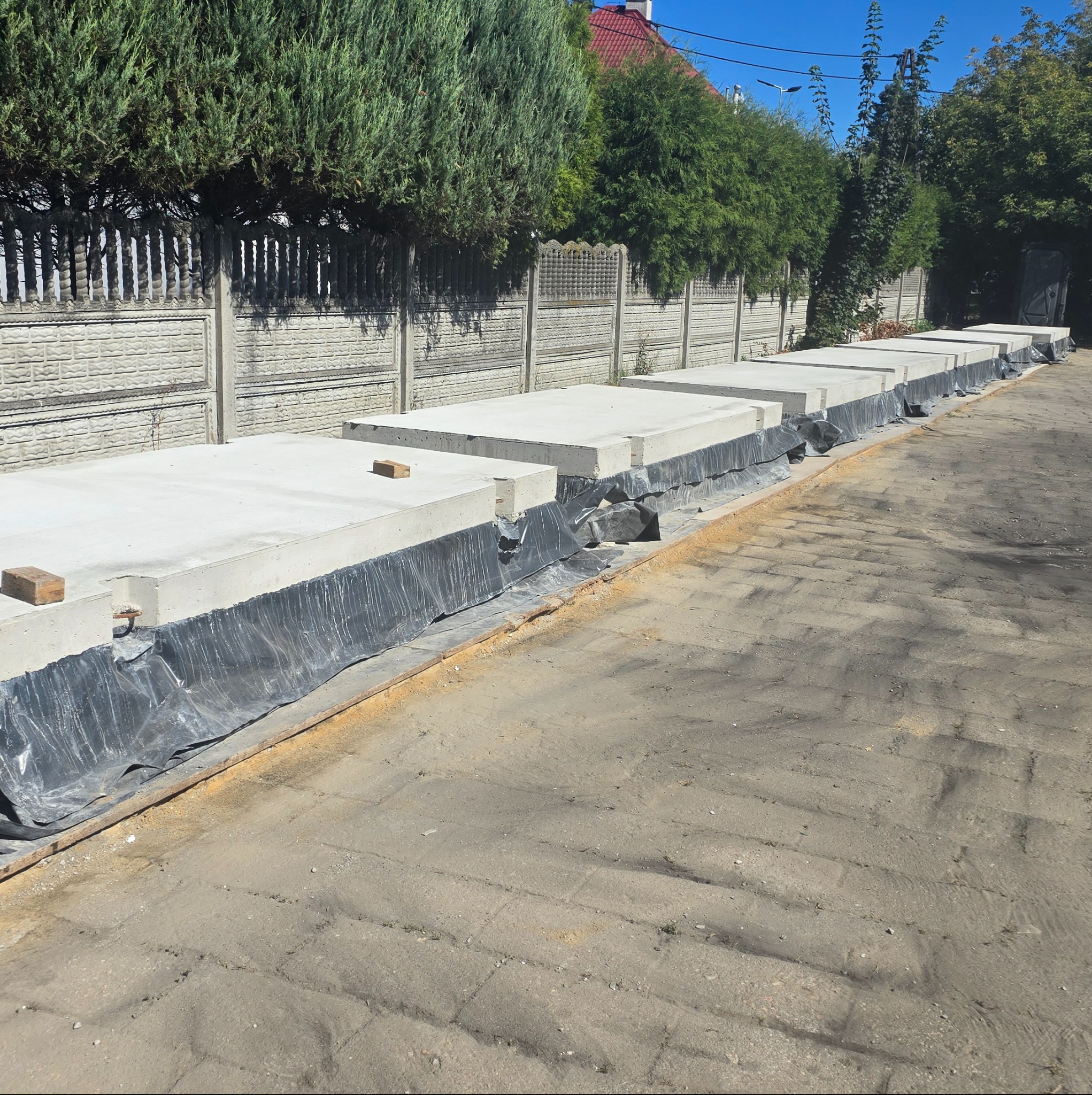 Ułożone płyty betonowe wzdłuż ogrodzenia, zabezpieczone folią. Widoczne elementy montażowe i drewniane podkładki. Jasne, naturalne światło dnia.