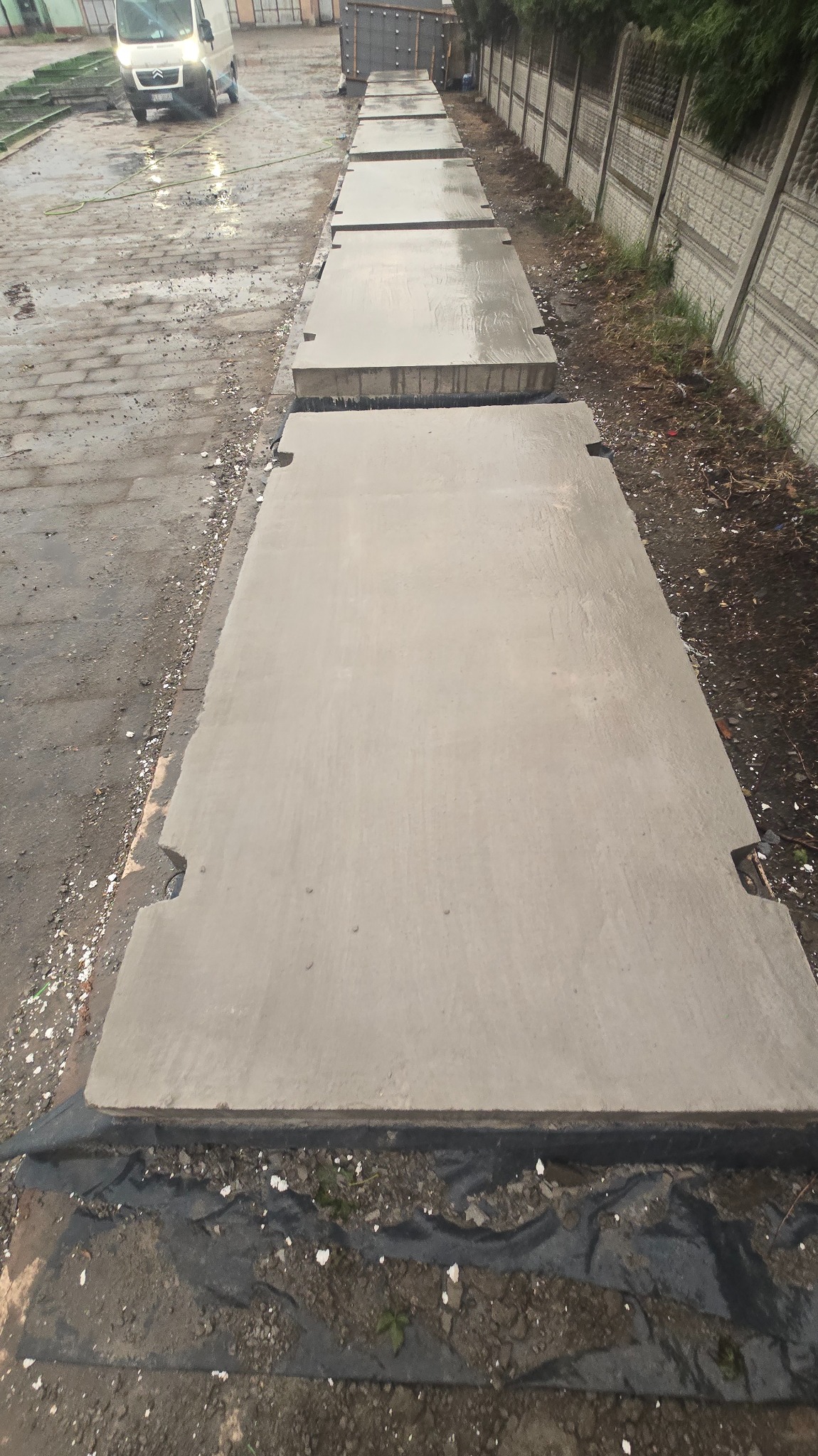 Ułożone płyty drogowe z betonu na mokrym podłożu, widoczne charakterystyczne wycięcia na krawędziach. W tle zaparkowany biały bus i betonowe ogrodzenie.