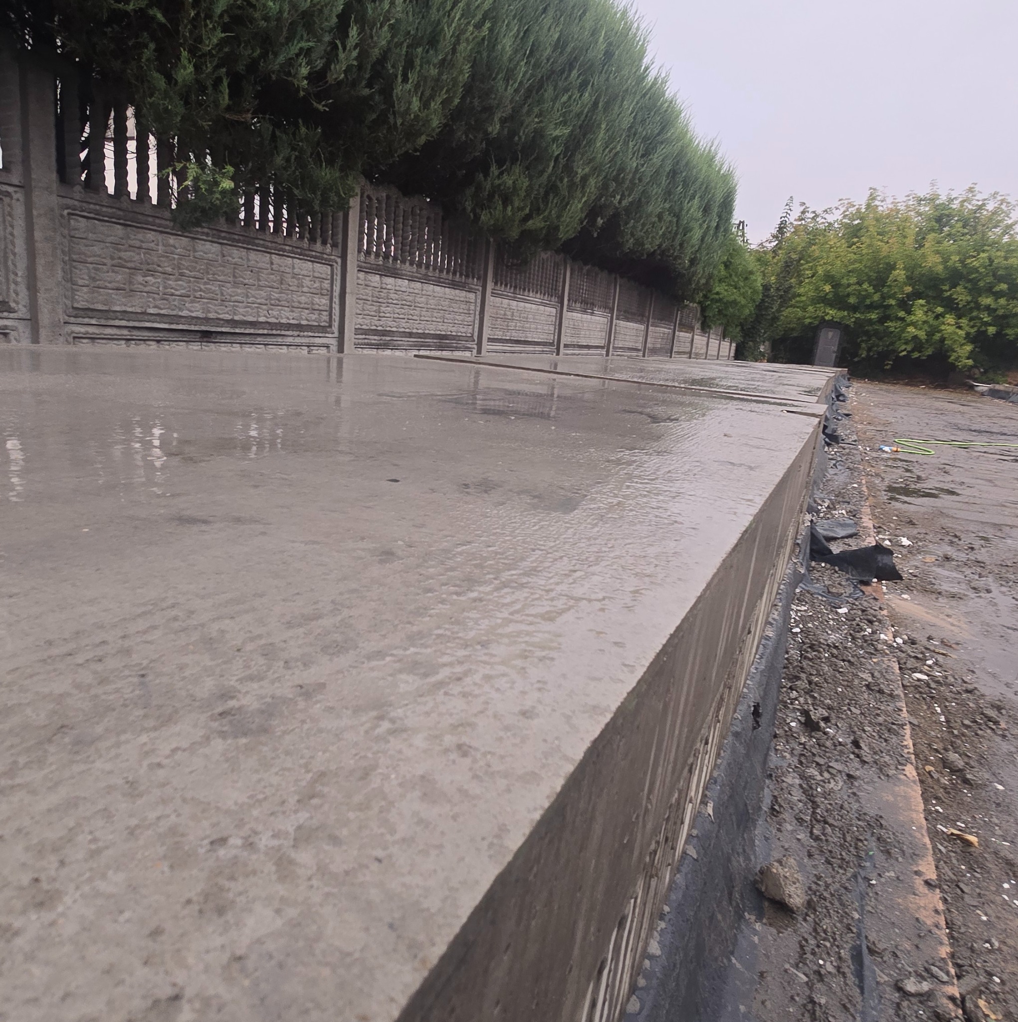 Ułożone, mokre płyty betonowe tworzące nawierzchnię, widoczne ogrodzenie z prefabrykatów betonowych i zieleń w tle. Ujęcie z niskiej perspektywy.