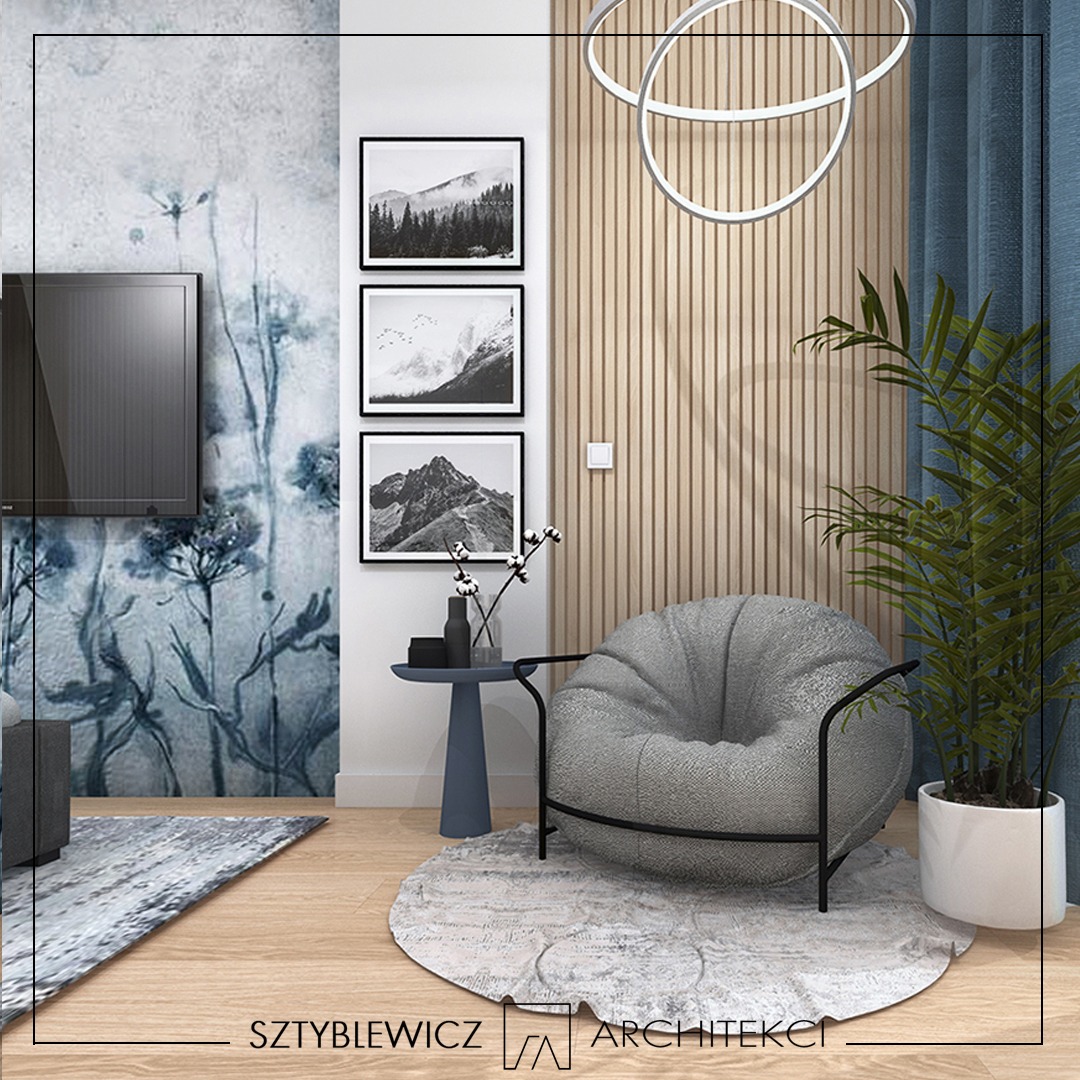 Stylowa aranżacja wnętrza z szarą pufą na okrągłym dywanie, drewnianą ścianą z oświetleniem i grafikami w czarnych ramkach. Roślina w białej donicy dodaje świeżości.