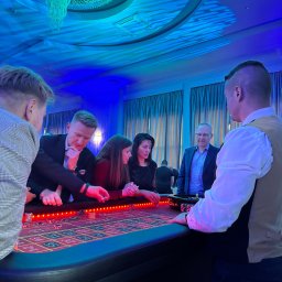 EVENTIVITY GROUP PAULINA TRYTKO - Uczestnicy imprezy firmowej grają w ruletkę przy stole kasyna, oświetlonym niebieskim światłem, w tle widoczny żyrandol i okna.