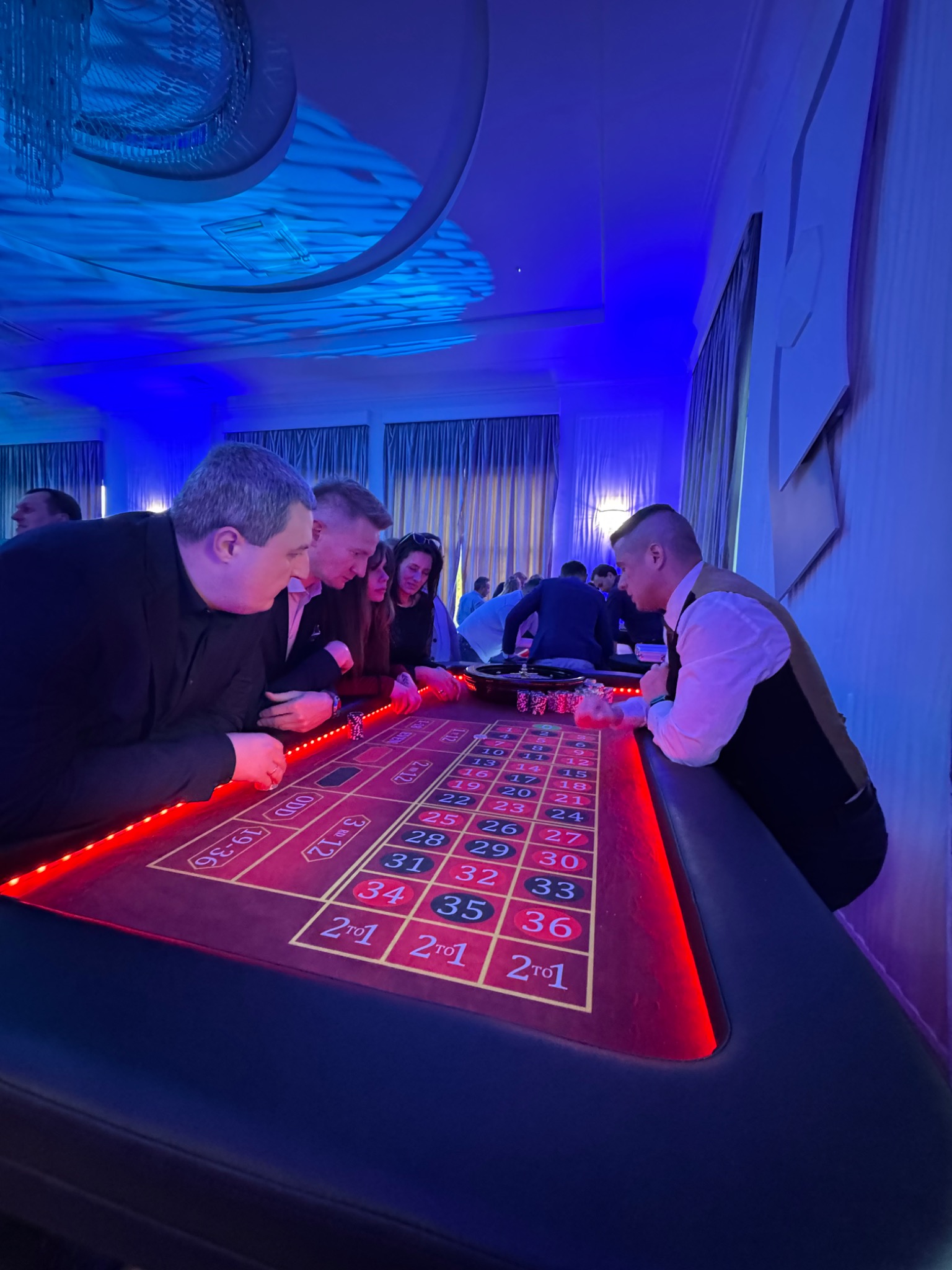 Stół do ruletki z czerwonym podświetleniem, przy którym stoją osoby w eleganckich strojach, w tle widoczne dekoracje sali w niebieskim oświetleniu.