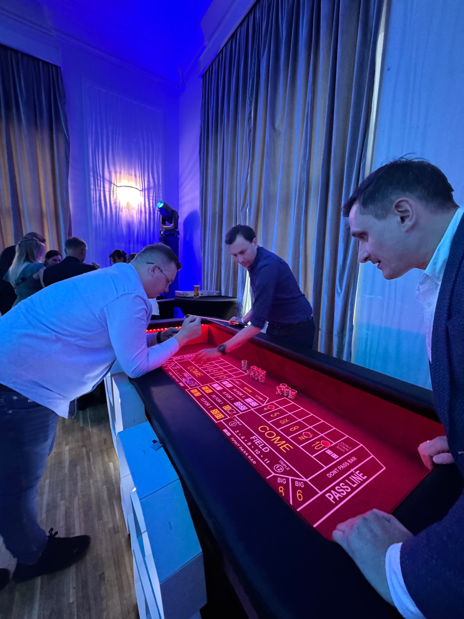 Obsługa stołu do gry w kości podczas imprezy. Trzech mężczyzn przy stole, w tle osoby i oświetlenie sceniczne. Kasyno na evencie firmowym.