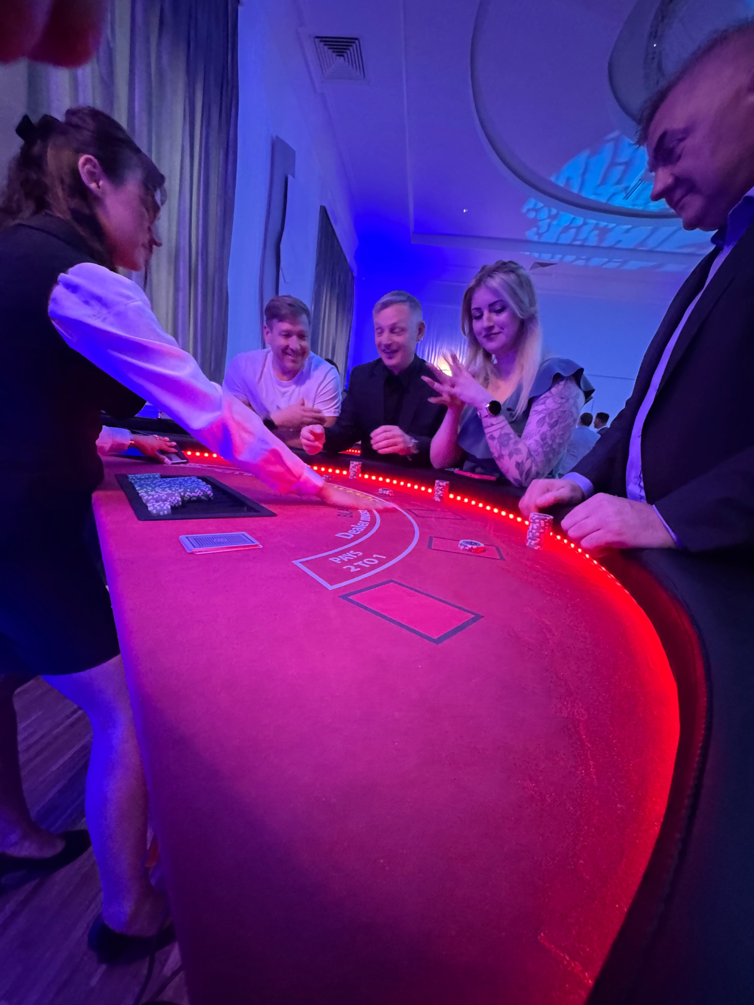 Stół do blackjacka z czerwonym obiciem, oświetlony neonowym światłem, wokół którego zgromadzeni są uśmiechnięci gracze i krupierka. Atmosfera kasyna na imprezie.