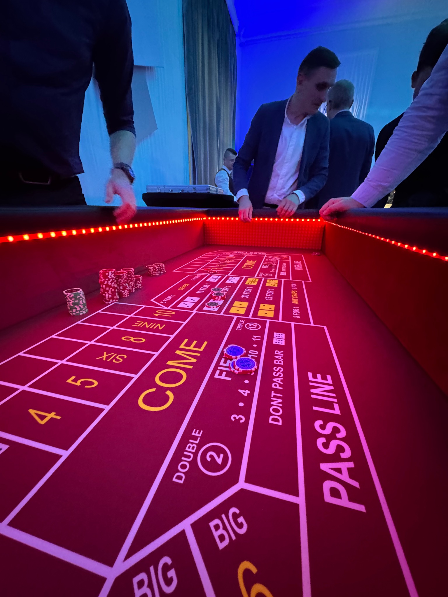 Stół do gry w kości z żetonami i graczami, oświetlony czerwonymi diodami LED, tworzący atmosferę kasyna na imprezie. Widok z perspektywy gracza.