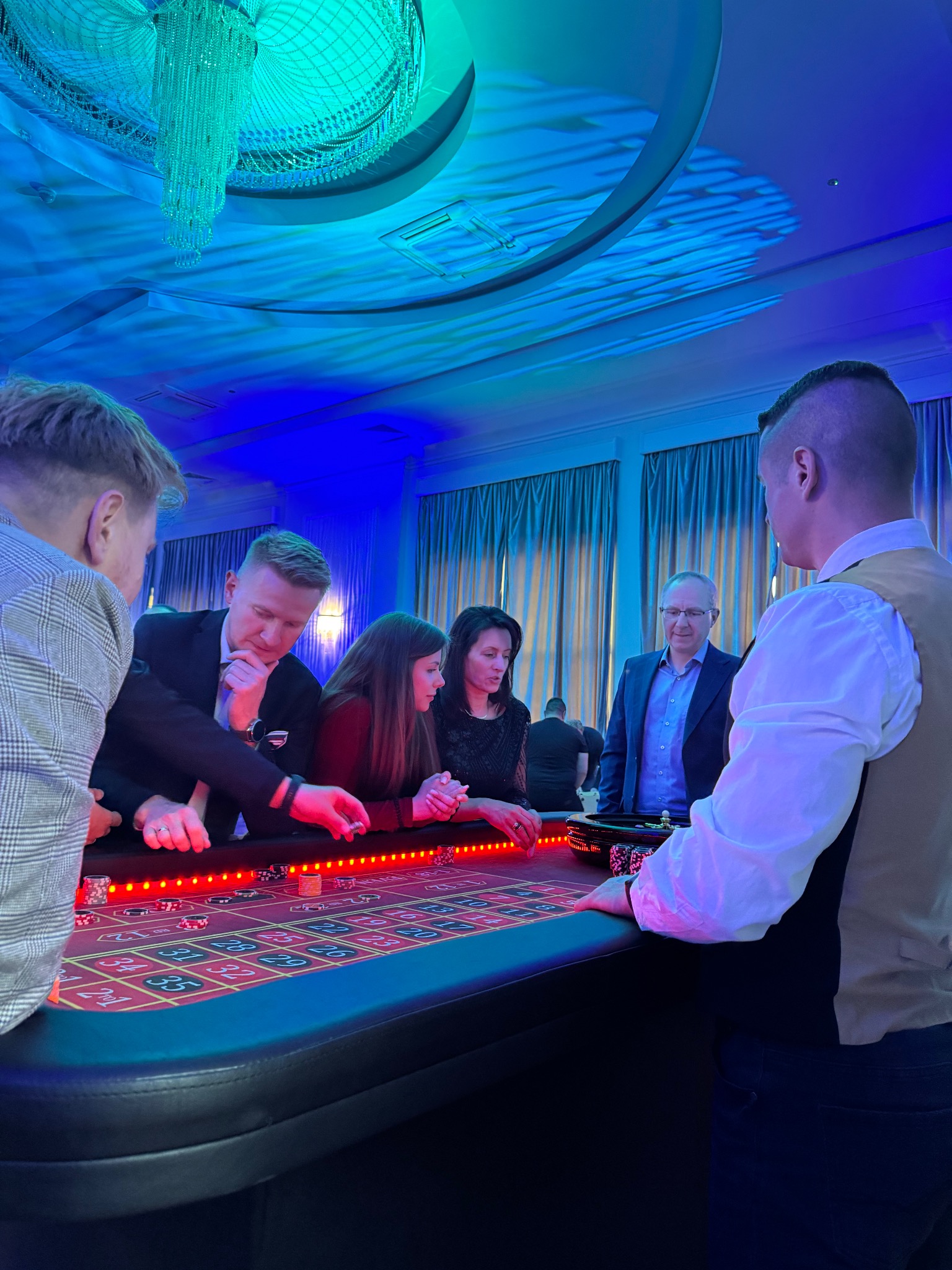 Uczestnicy imprezy firmowej grają w ruletkę przy stole kasyna, oświetlonym niebieskim światłem, w tle widoczny żyrandol i okna.