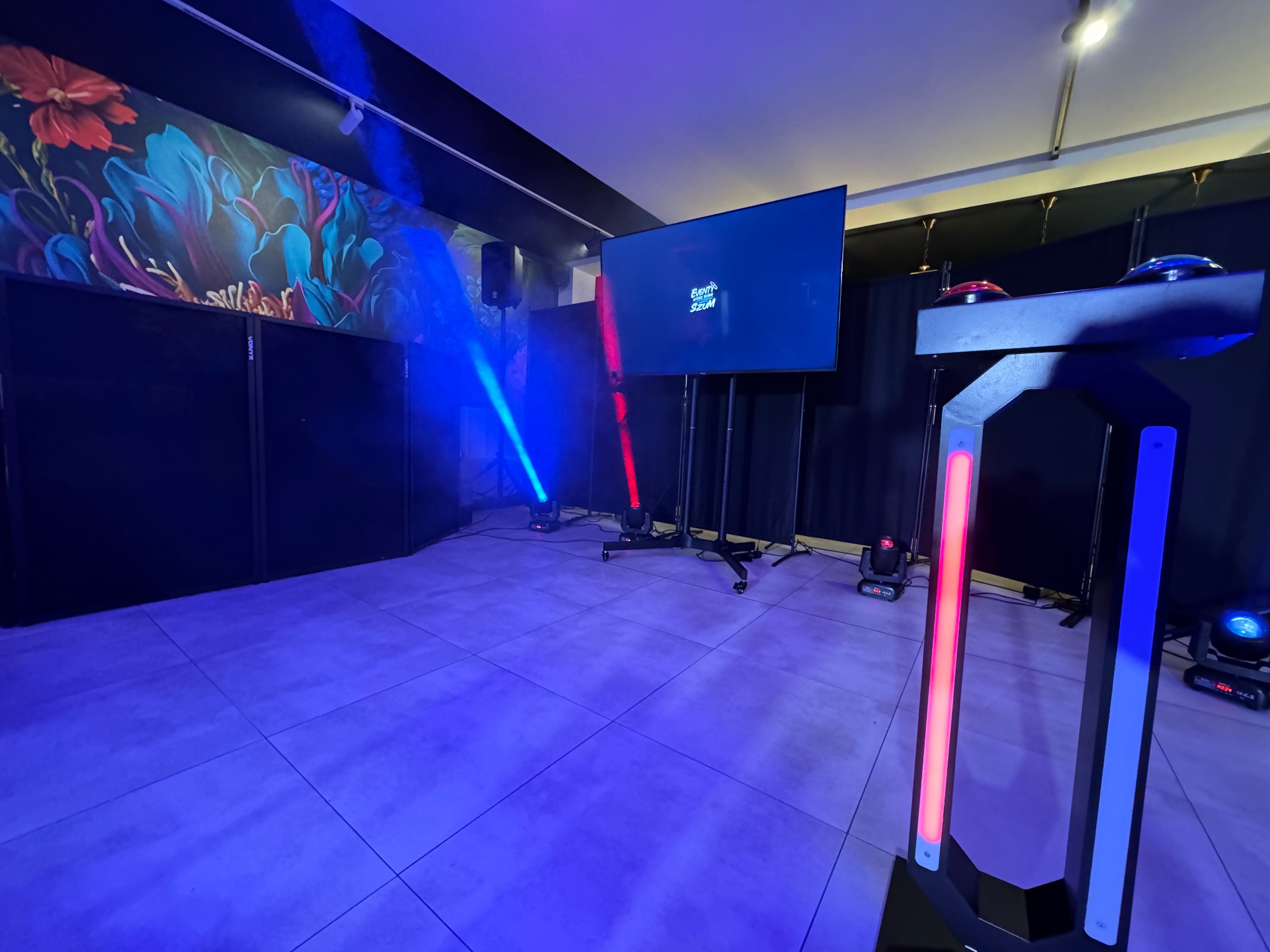 Nowoczesna fotobudka z ekranem, oświetleniem LED (czerwone i niebieskie) i interaktywnymi przyciskami, ustawiona w pomieszczeniu z dekoracyjną tapetą i profesjonalnym nagłośnieniem.