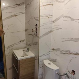 PRZEMYSŁAW WĘDZIKOWSKI DZIK BUD - Wykończona łazienka z białymi płytkami imitującymi marmur, drewnianą szafką pod umywalką i toaletą. Widoczne przyłącza wodne. Minimalistyczny styl.