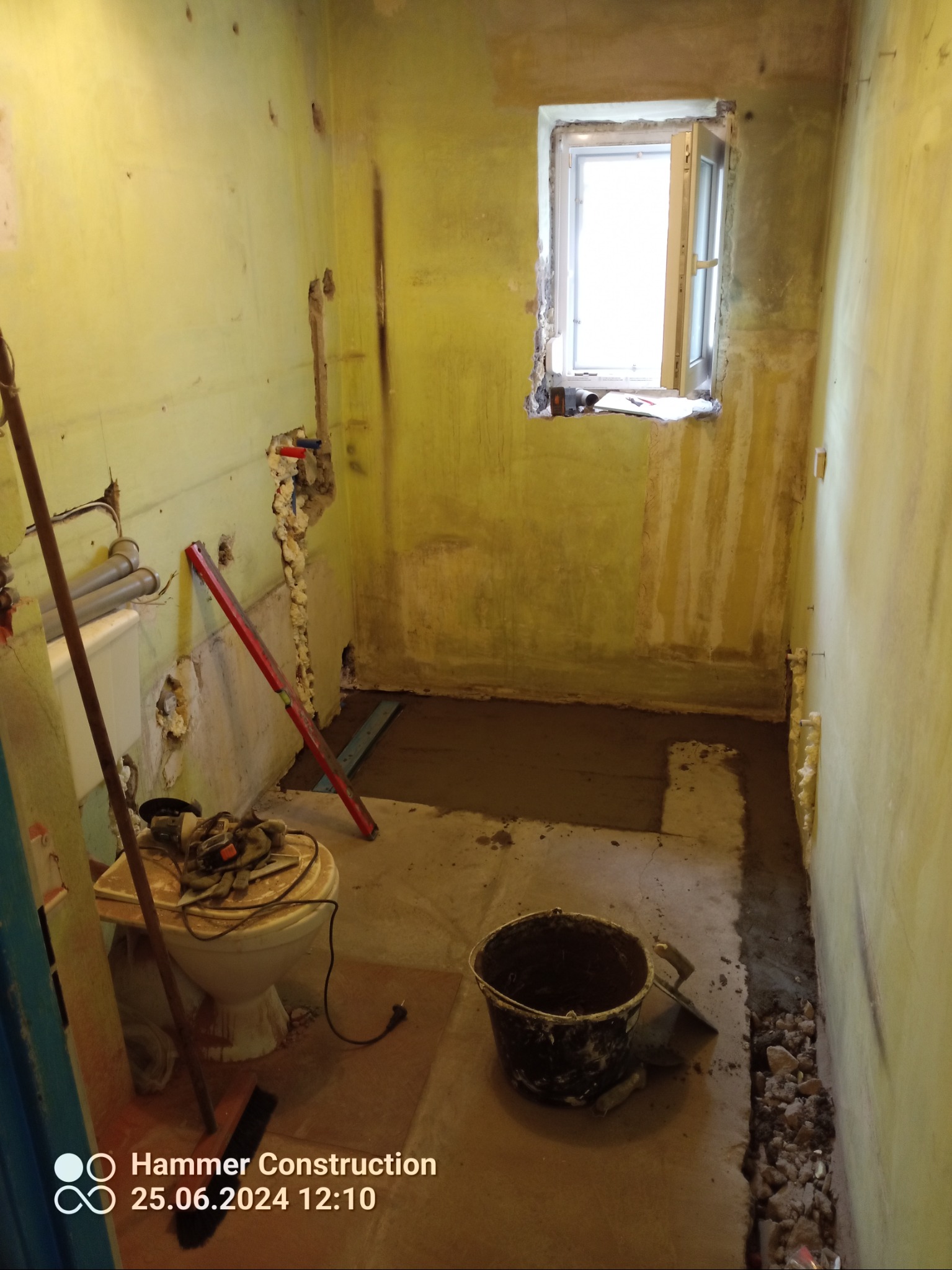 Wnętrze łazienki w trakcie prac przygotowawczych do remontu. Widoczne ściany z usuniętymi fragmentami tynku, częściowo wylana wylewka, narzędzia i wiadro.