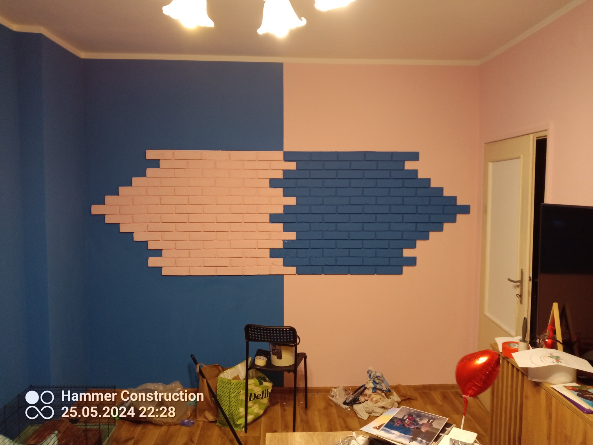 Dwukolorowa ściana z dekoracyjną cegłą w odcieniach różu i niebieskiego, tworząca unikalny wzór. Widoczne niedoskonałości i bałagan na podłodze.