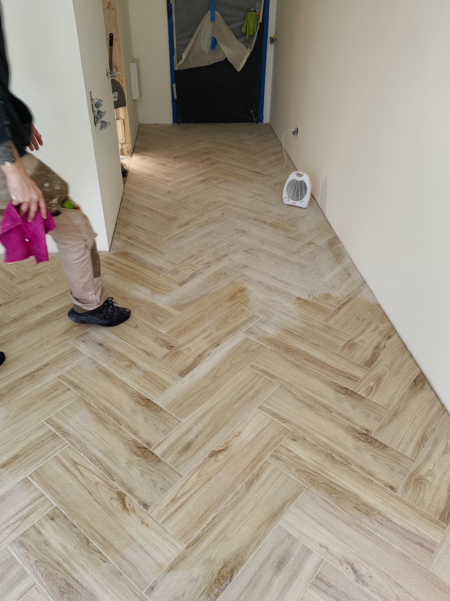 Ułożona podłoga z płytek imitujących drewno w jodełkę, widoczne fragmenty osoby czyszczącej podłogę, w tle drzwi zabezpieczone folią malarską i grzejnik.