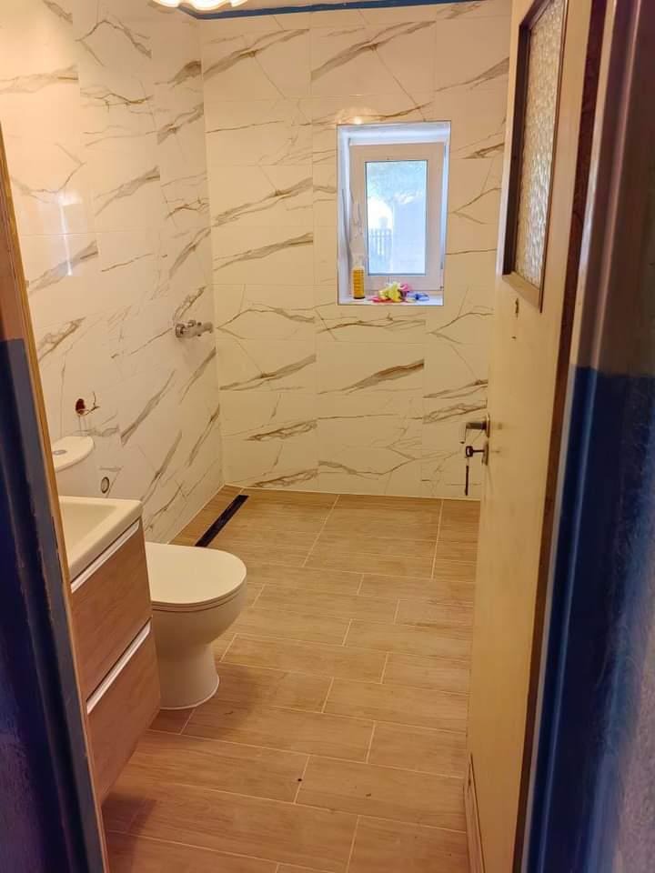 Jasna łazienka po remoncie: marmurowe płytki ścienne, drewnopodobna podłoga, okno z widokiem na zieleń. Umywalka z szafką i toaleta w neutralnych barwach.