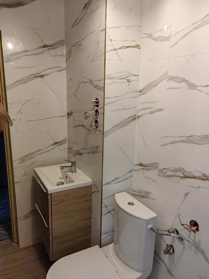 Wykończona łazienka z białymi płytkami imitującymi marmur, drewnianą szafką pod umywalką i toaletą. Widoczne przyłącza wodne. Minimalistyczny styl.