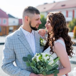 Daria Chaika - Uśmiechnięta para młoda w plenerze: on w szarej marynarce w kratę, ona w białej sukni z bukietem. W tle budynek z czerwoną dachówką i fontanna.