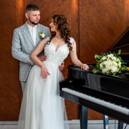 Daria Chaika - Elegancka para młoda w jasnych strojach pozuje przy czarnym fortepianie na tle drewnianej ściany. Bukiet białych róż leży na instrumencie. Uśmiechnięci patrzą na siebie.