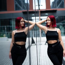 Daria Chaika - Kobieta z czerwonymi włosami w czarnym stroju pozuje, opierając się o lustrzaną ścianę budynku. Widoczne odbicie, miejski krajobraz w tle.
