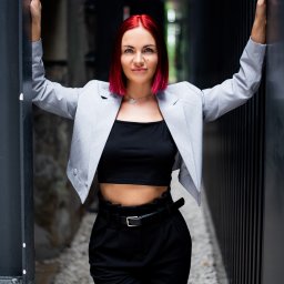 Daria Chaika - Kobieta z czerwonymi włosami w szarym żakiecie i czarnym topie, oparta o czarne metalowe elementy architektoniczne. Uśmiech, naturalne światło.