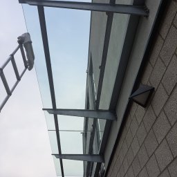 Andrzej Sładkowski - Szklany daszek nad wejściem do budynku, widoczna drabina z szmatką, sugerująca niedawne mycie. Ujęcie z dołu, podkreślające konstrukcję i czystość szkła.