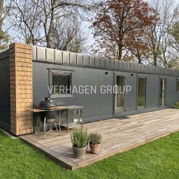 Verhagen Kraków Sp. z o. o. - Gotowy, szary dom modułowy z drewnianą elewacją i tarasem, grill na zewnątrz. Widok z poziomu trawnika, drzewa w tle. Nowoczesna architektura.