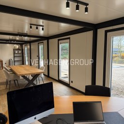 Verhagen Kraków Sp. z o. o. - Wnętrze modułowego biura z drewnianym stołem konferencyjnym, oświetleniem punktowym i widokiem na zewnątrz przez okna. Na pierwszym planie biurko z komputerami.