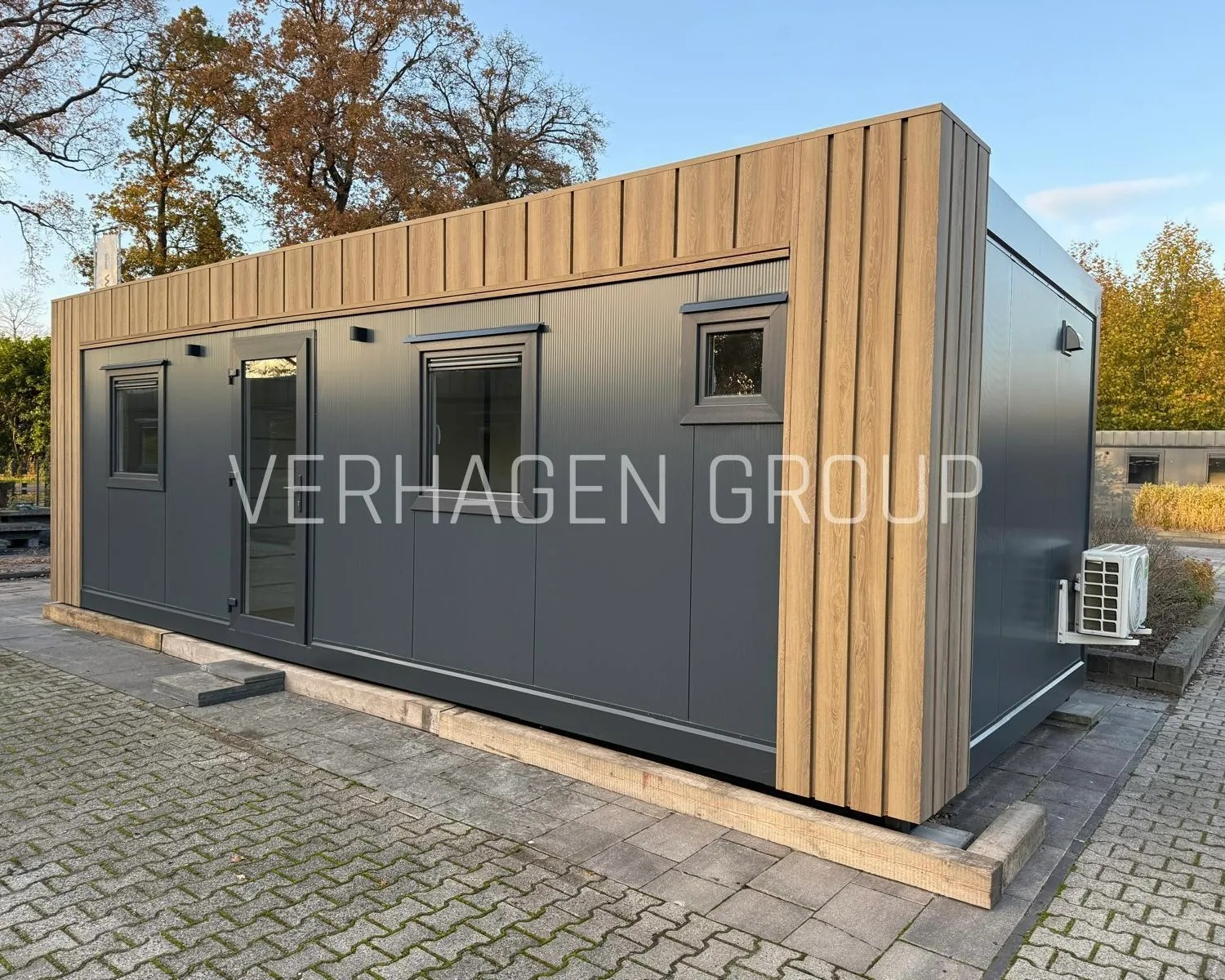 Nowoczesny, szary pawilon z drewnianymi akcentami i klimatyzacją, ustawiony na kostce brukowej. Widoczne okna, drzwi i logo firmy na elewacji.