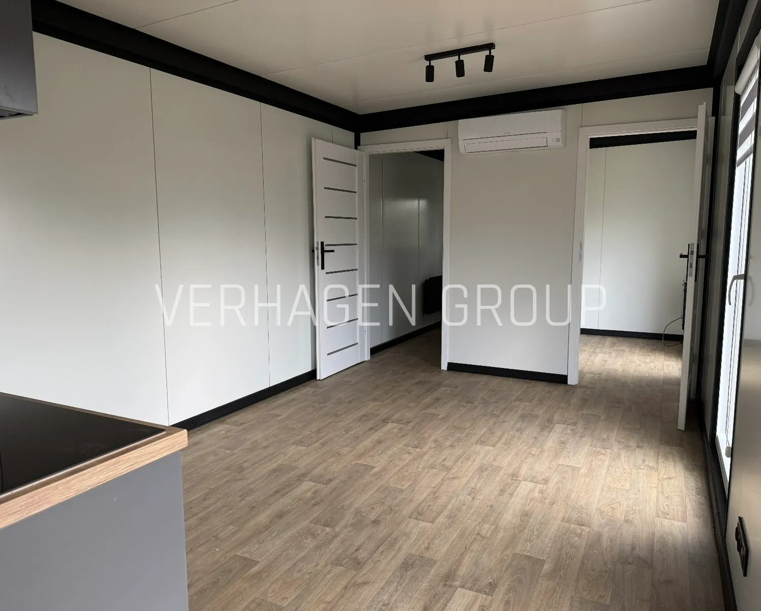 Wnętrze modułowego domu z drewnianą podłogą, białymi ścianami i czarnymi akcentami. Widoczne drzwi, klimatyzacja i oświetlenie sufitowe.