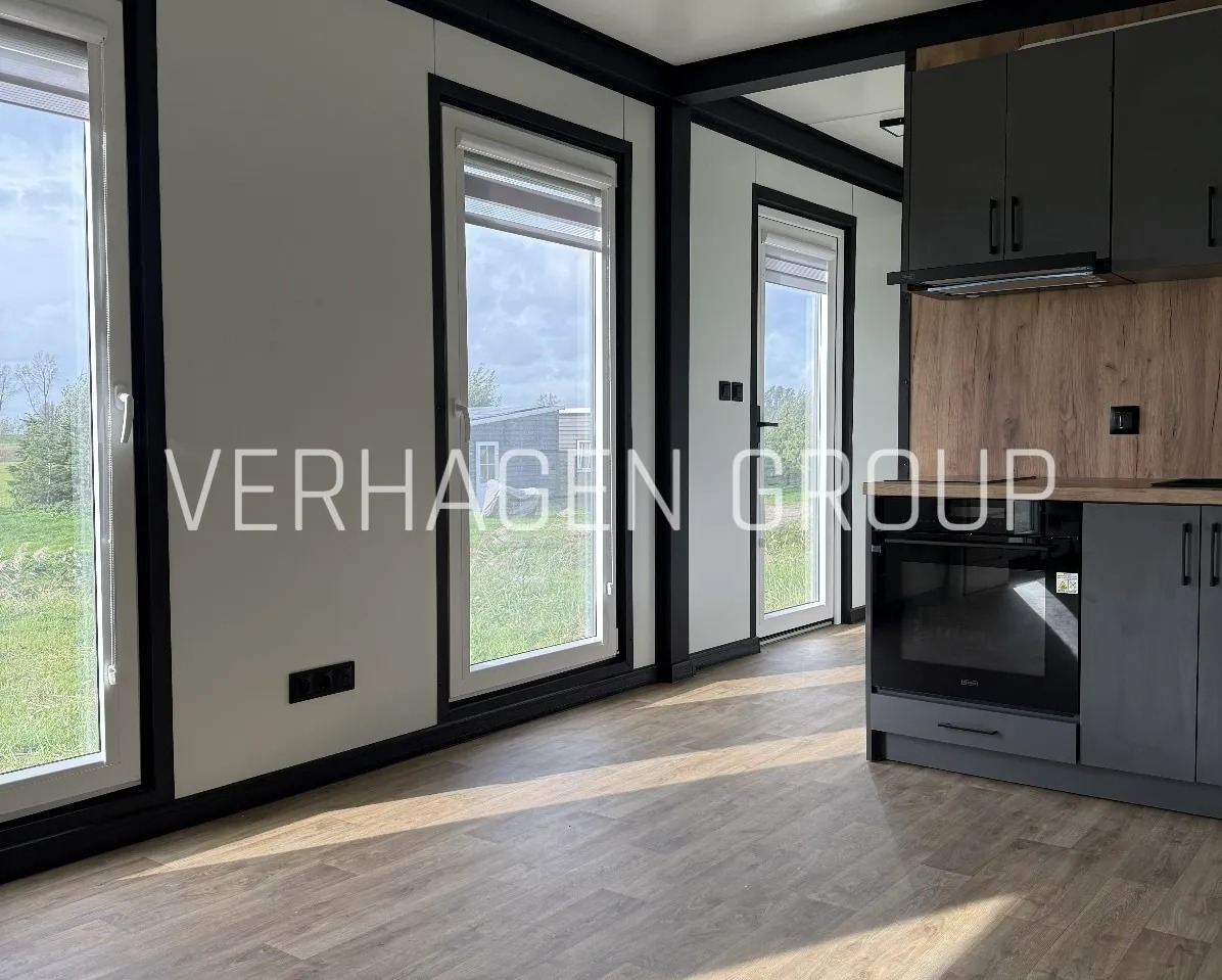 Jasne wnętrze z oknami i szarą zabudową kuchenną. Widok na zewnątrz przez okna. Podłoga imitująca drewno. Minimalistyczny design.