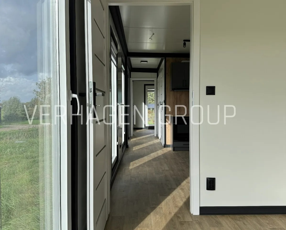 Wnętrze modułowego domu z widokiem na zewnątrz przez duże okna. Jasne ściany, drewniana podłoga i minimalistyczny design. Widoczna kuchnia w tle.