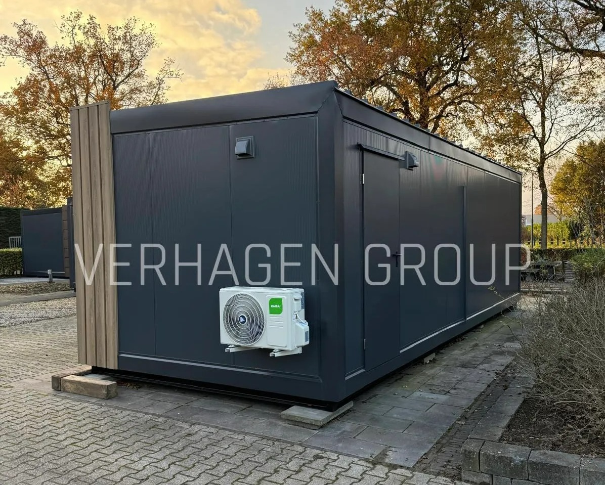 Szary budynek modułowy z klimatyzacją i drewnianymi panelami, otoczony zielenią i kostką brukową, w jesiennej scenerii. Widoczny fragment innego budynku.