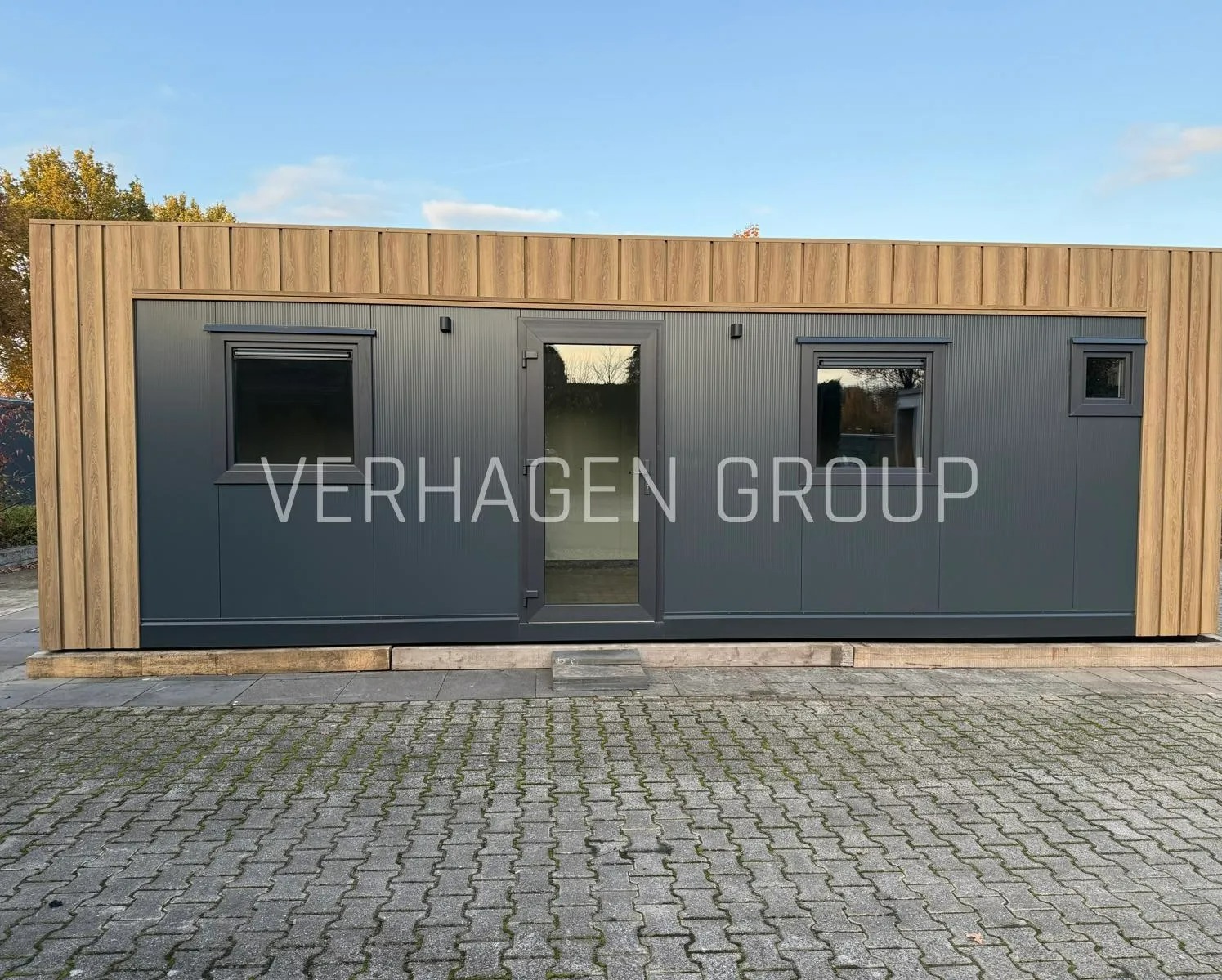Nowoczesny, szary budynek z drewnianymi akcentami i małymi oknami, ustawiony na kostce brukowej. Widoczne logo firmy na elewacji. Minimalistyczny design.