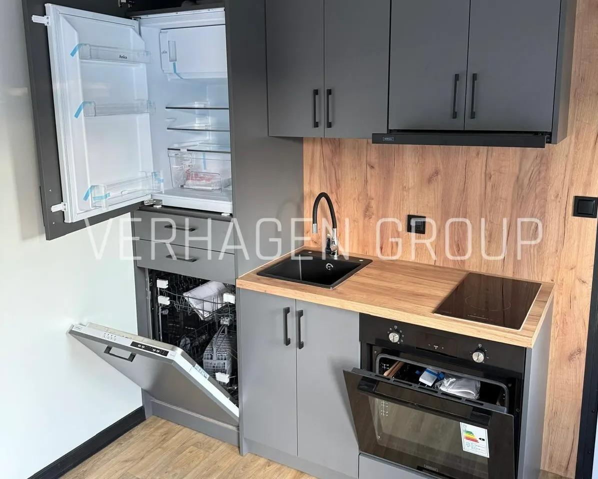 Szara, kompaktowa kuchnia w zabudowie: lodówka, zmywarka, piekarnik i płyta indukcyjna. Drewnopodobny panel ścienny. Idealna do małych przestrzeni.