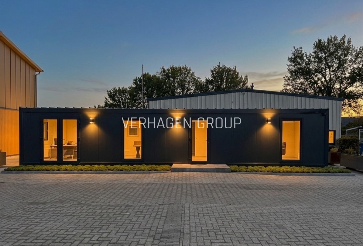 Nowoczesny, czarny dom modułowy z oświetleniem zewnętrznym i dużymi oknami, prezentujący minimalistyczny design i funkcjonalność. Widoczny napis 'VERHAGEN GROUP'.