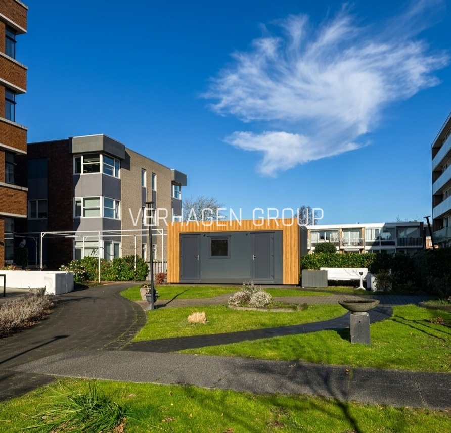 Nowoczesny, parterowy dom modułowy z szarymi drzwiami i oknem, otoczony zielenią i budynkami mieszkalnymi w słoneczny dzień. Widok z poziomu gruntu.