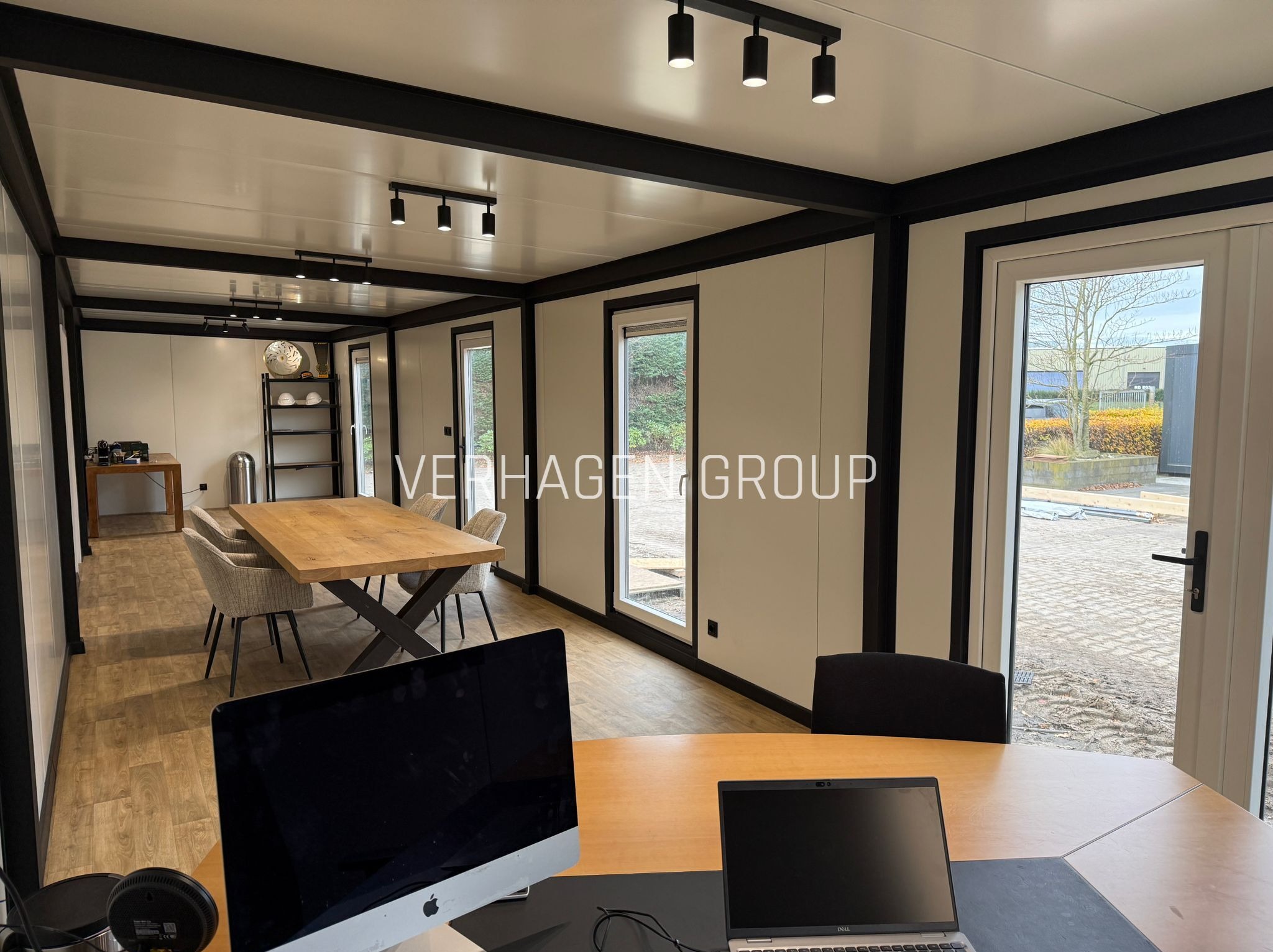 Wnętrze modułowego biura z drewnianym stołem konferencyjnym, oświetleniem punktowym i widokiem na zewnątrz przez okna. Na pierwszym planie biurko z komputerami.