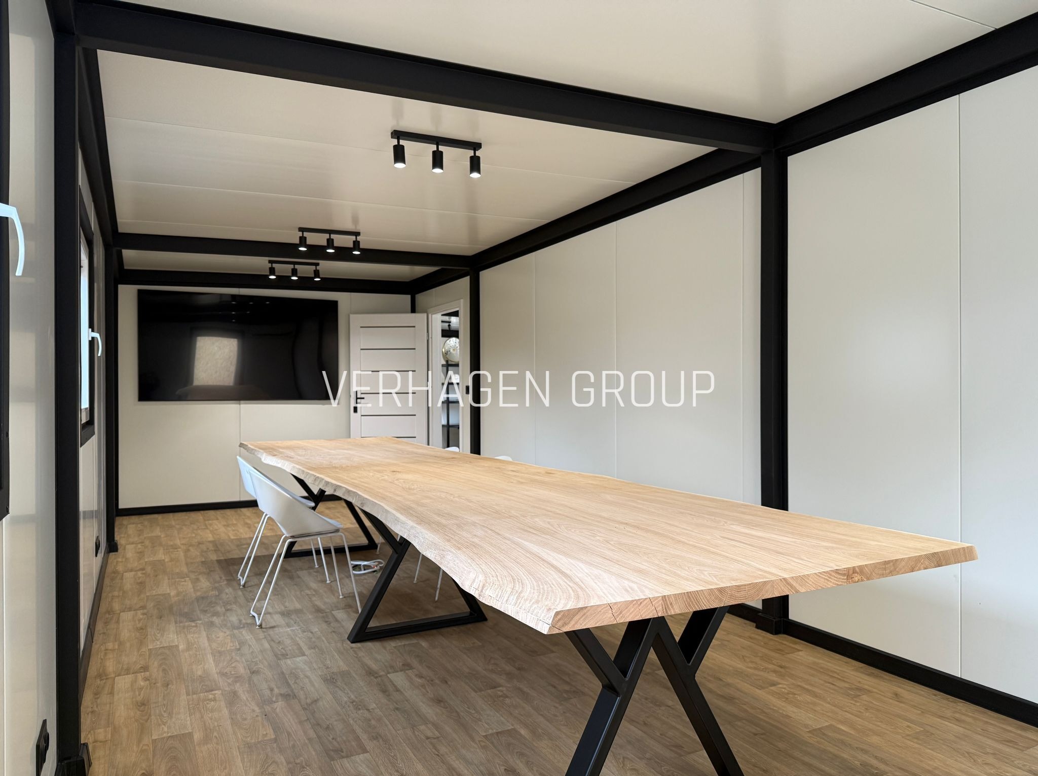 Wnętrze modułowego biura z dużym, drewnianym stołem na metalowych nogach. Czarne ramy konstrukcyjne, białe ściany i podłoga imitująca drewno. Nowoczesne oświetlenie.