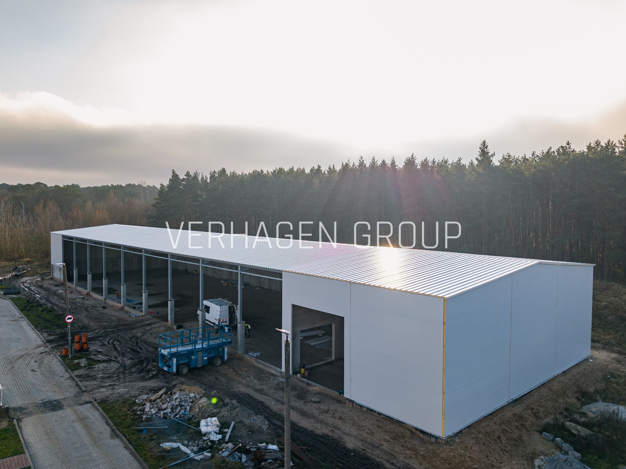 Nowoczesna hala stalowa z białymi ścianami i srebrnym dachem, widok z lotu ptaka, na tle lasu i pochmurnego nieba. Widoczny podnośnik koszowy i częściowo wykończony teren.