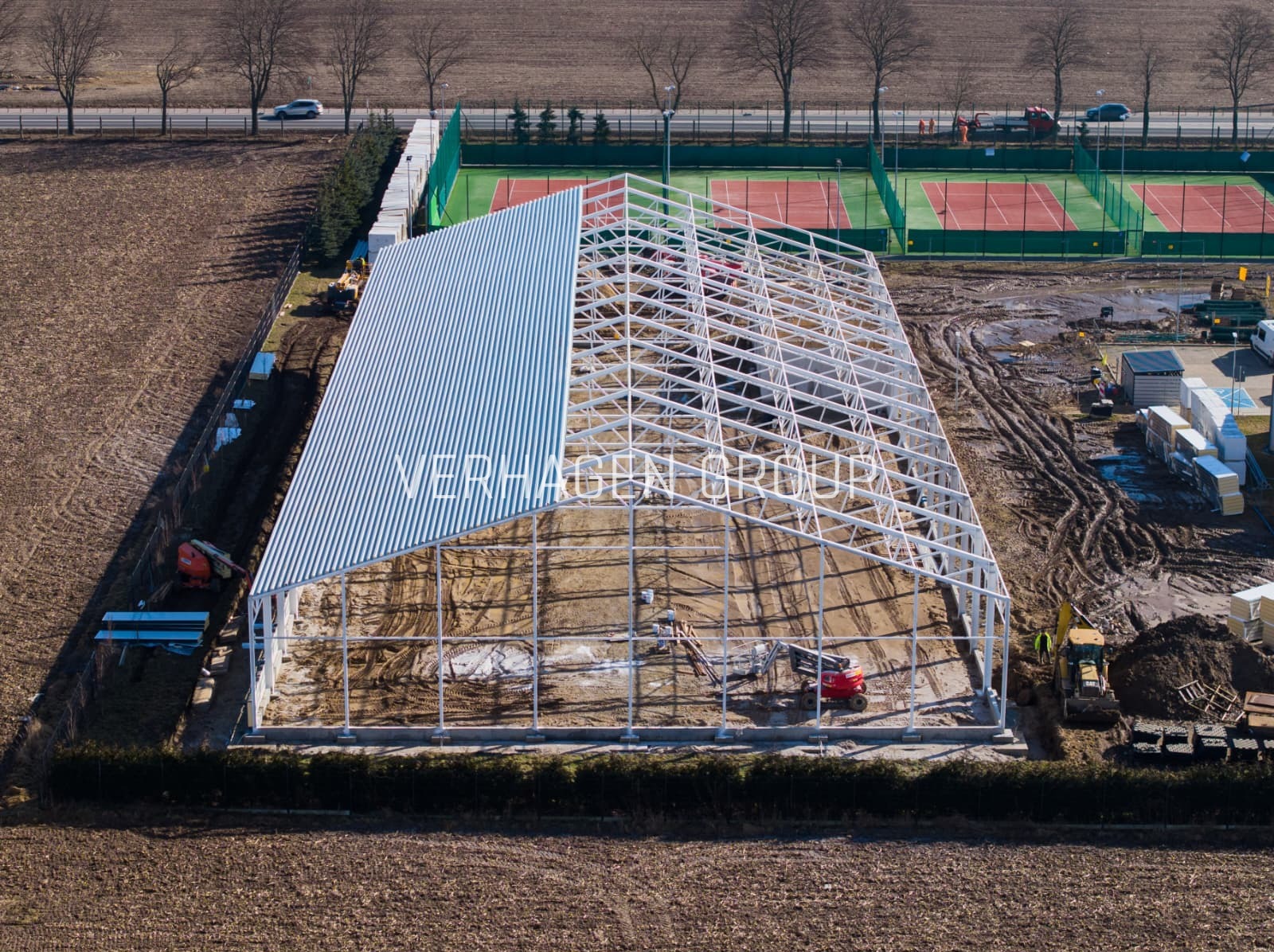 Budowa hali z częściowo ukończonym dachem z blachy falistej, widok z góry na plac budowy z maszynami i kortami tenisowymi w tle. Konstrukcja z logo firmy.