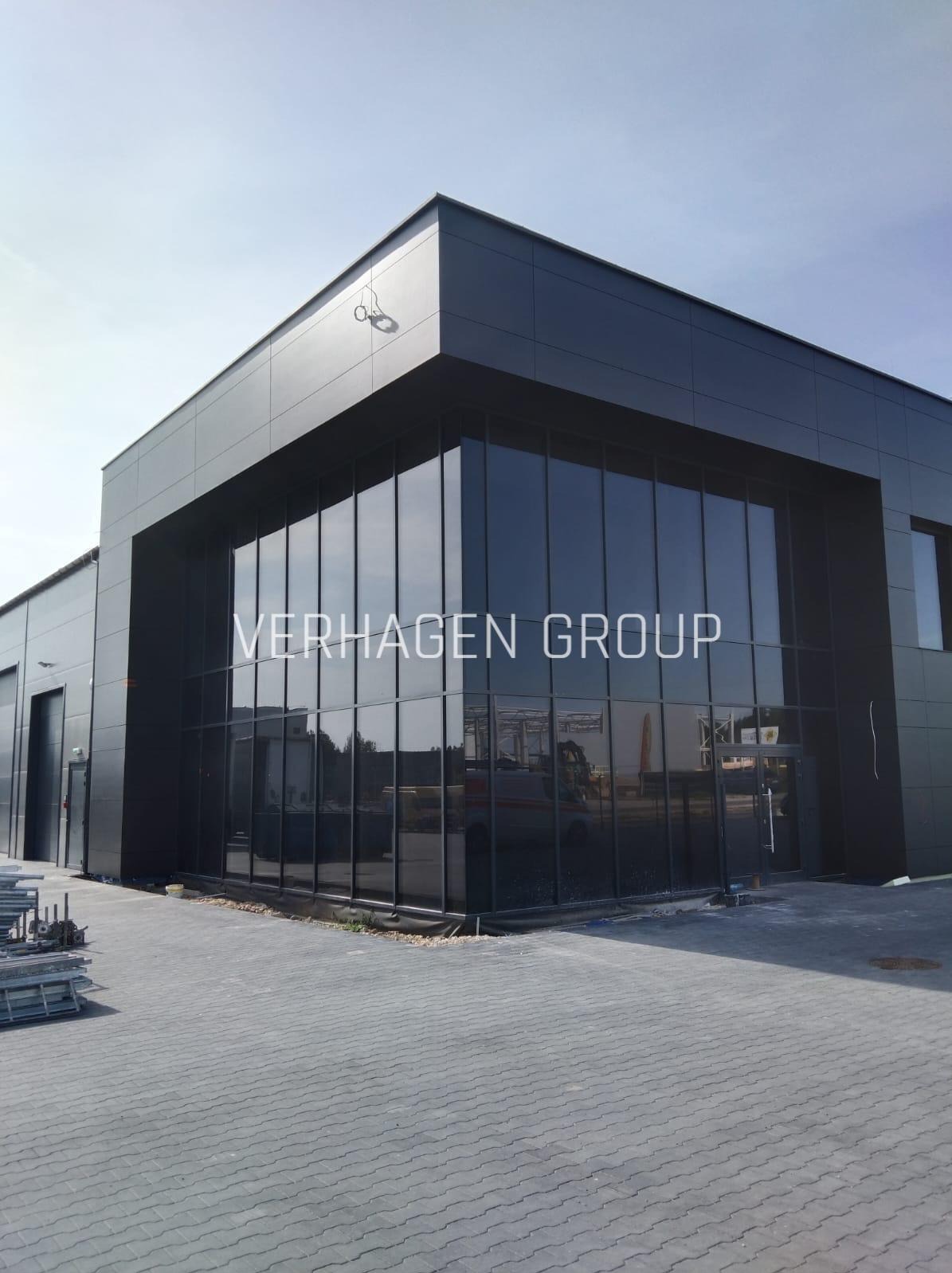 Nowoczesna hala przemysłowa z czarną elewacją i dużymi oknami, na których widnieje napis 'VERHAGEN GROUP'. Na pierwszym planie kostka brukowa i elementy konstrukcyjne.