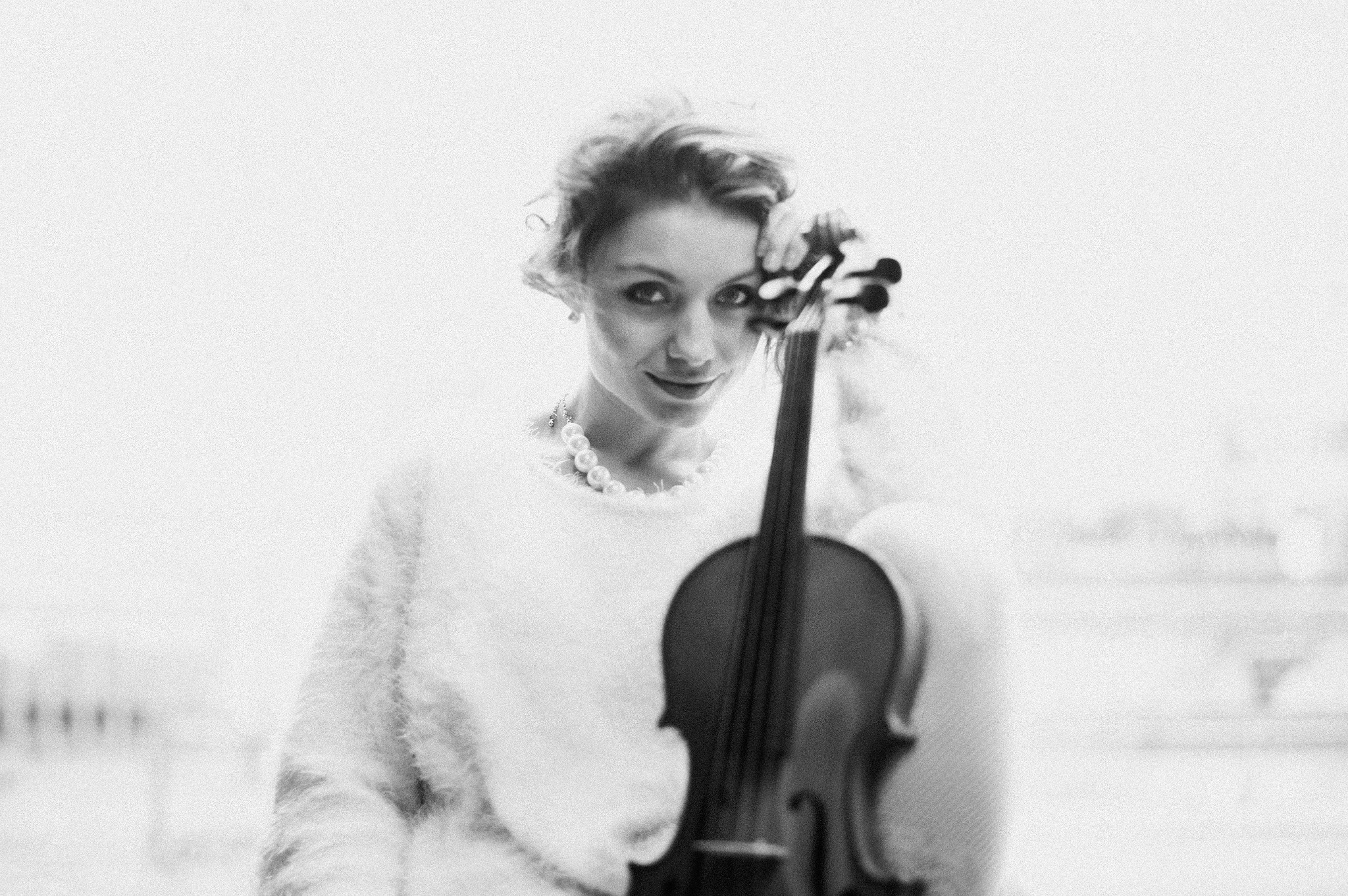 Portret kobiety z kręconymi włosami i perłami, trzymającej skrzypce na rozmytym tle. Zdjęcie czarno-białe, z delikatnym uśmiechem i spojrzeniem w obiektyw.