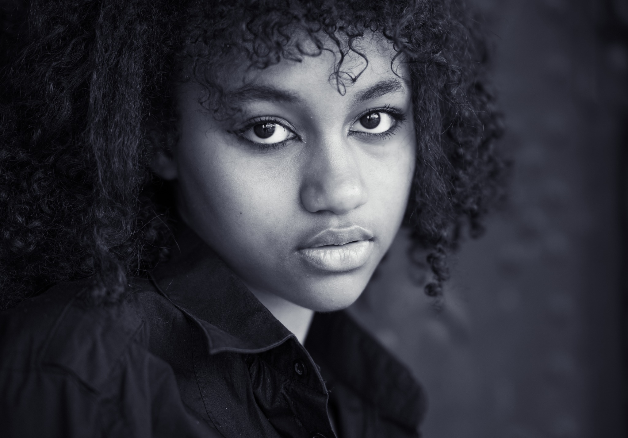 Monochromatyczny portret młodej kobiety z kręconymi włosami, ubrana w ciemną koszulę, z delikatnym makijażem i intensywnym spojrzeniem skierowanym wprost w obiektyw.