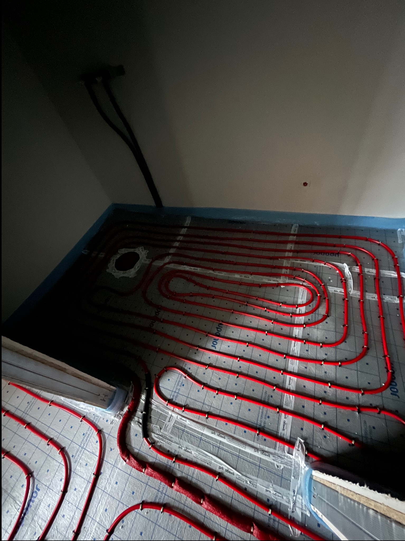 Czerwone rury ogrzewania podłogowego ułożone na izolacji z widocznymi mocowaniami. Fragment instalacji przy ścianie z przewodami. Ujęcie z góry, w trakcie montażu.