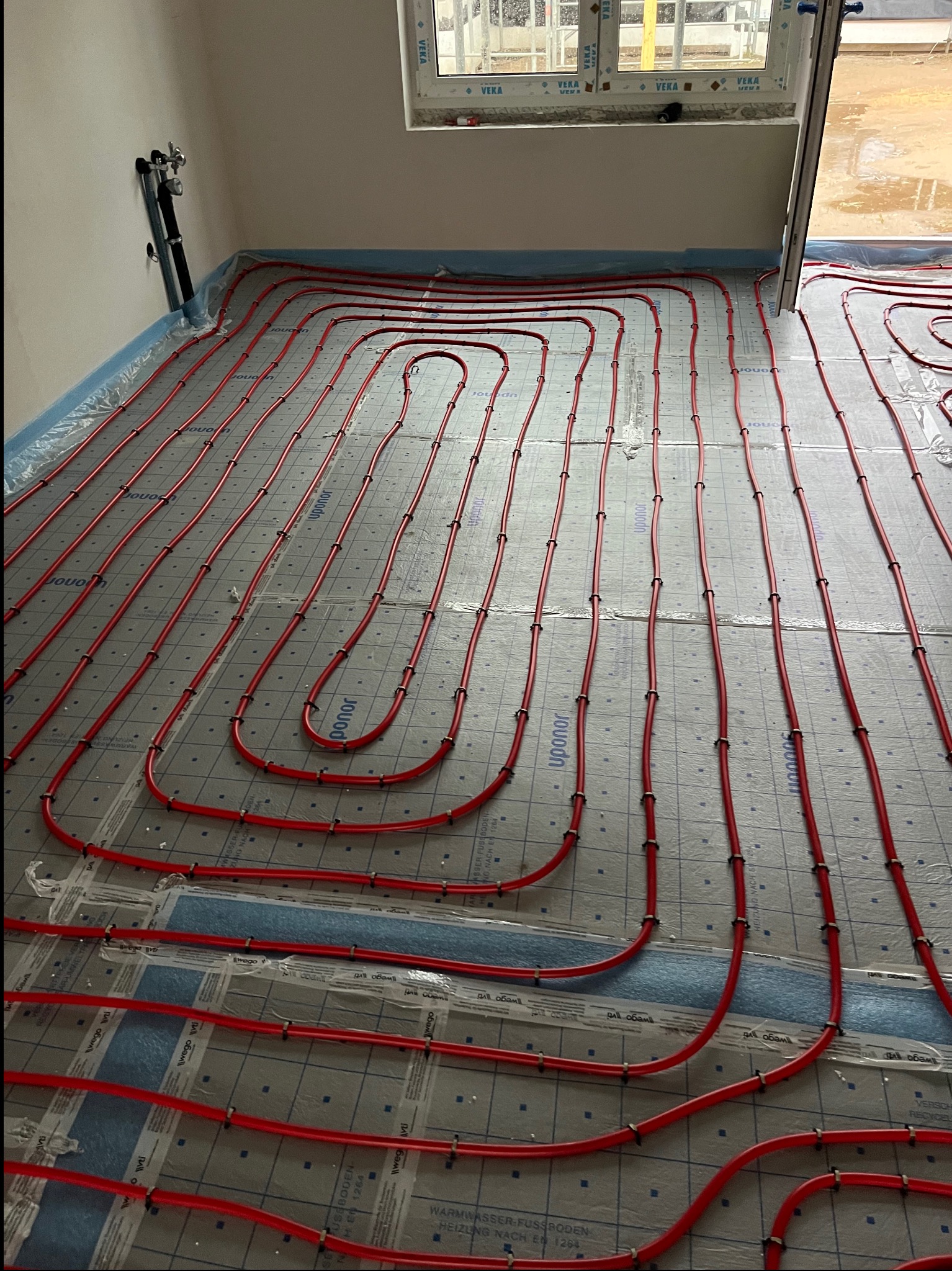 Czerwone rury ogrzewania podłogowego ułożone na izolacji z folią aluminiową, widoczne mocowania. Instalacja przed wylaniem posadzki, okno w tle.