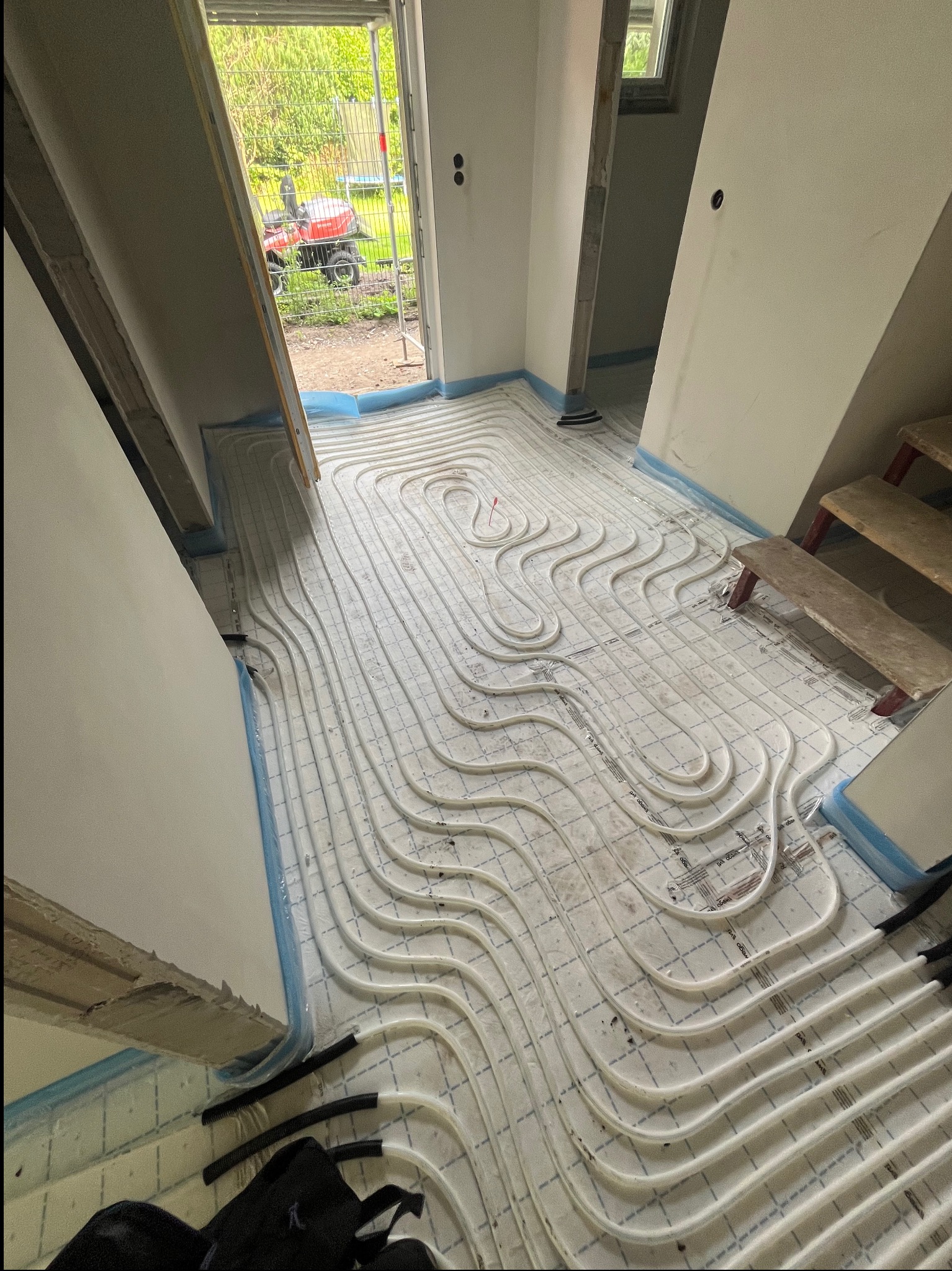 Białe rury ogrzewania podłogowego ułożone na podkładzie z folią, widoczne fragmenty ścian i schodów, przez otwarte drzwi widać traktor na zewnątrz.
