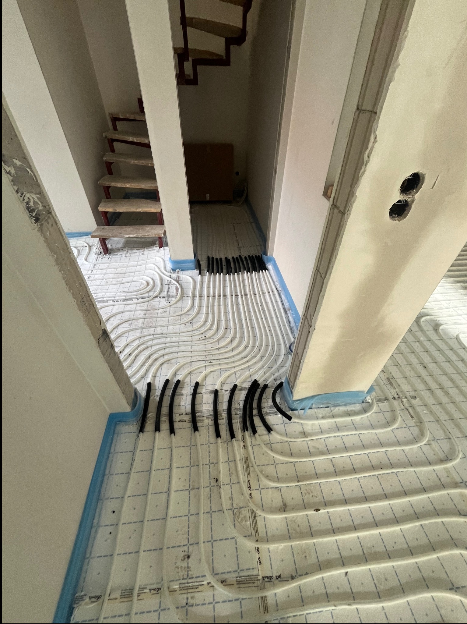 Białe rurki instalacji podłogowej ułożone na siatce, widoczne czarne elementy mocujące, niebieska folia izolacyjna przy ścianach, surowe ściany i schody w tle.