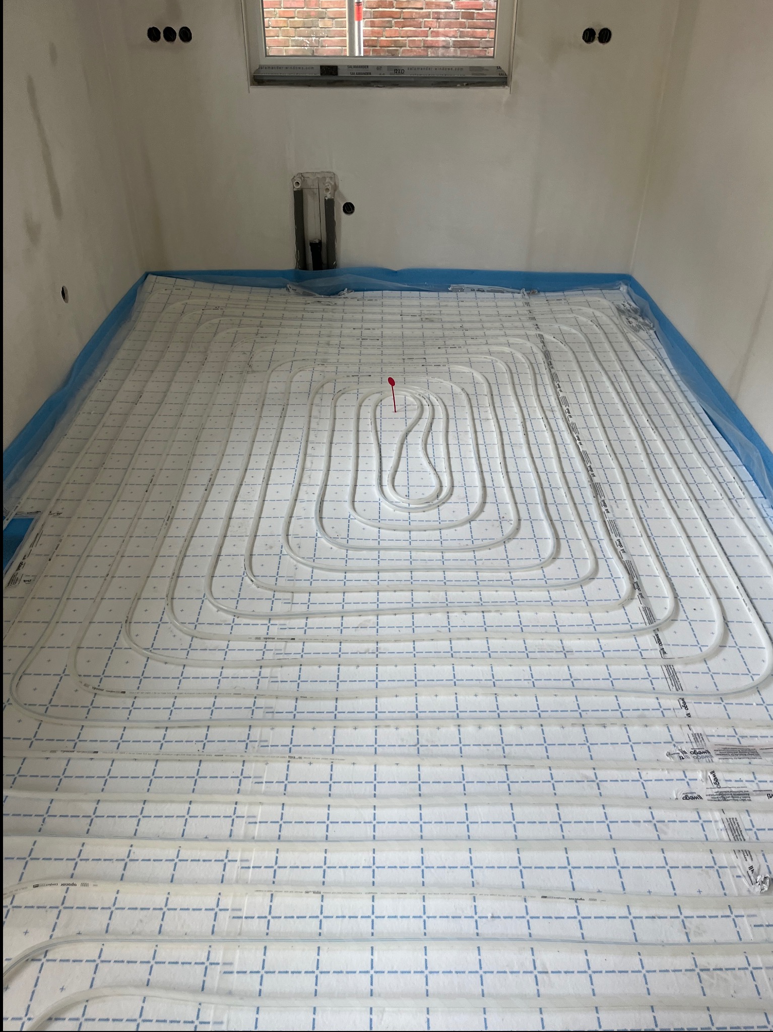 Białe rury ogrzewania podłogowego ułożone na izolacji z niebieską siatką w surowym wnętrzu. Widoczne otwory w ścianie i okno z ceglanym murem w tle.