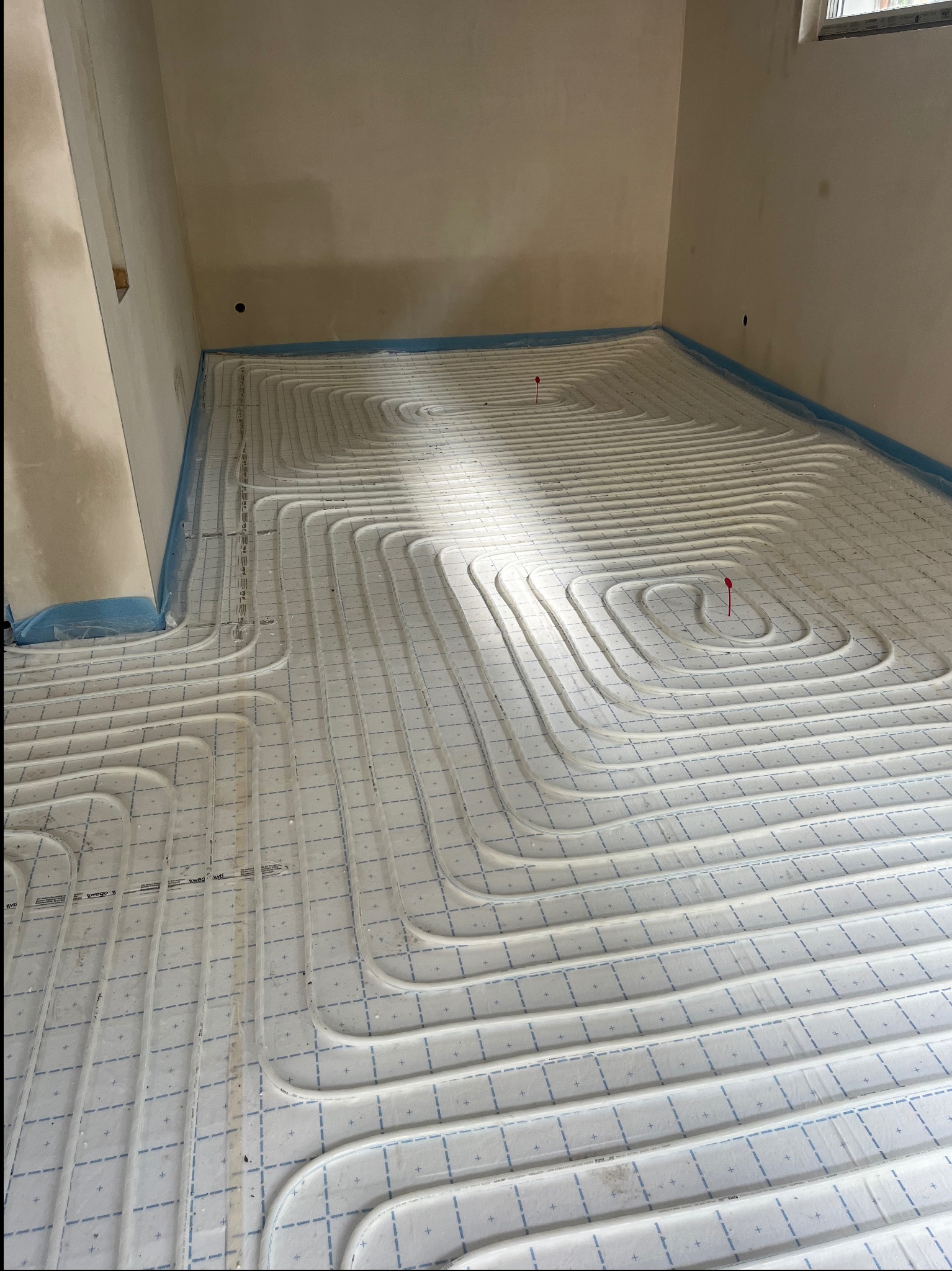 Białe rury ogrzewania podłogowego ułożone spiralnie na izolacji z niebieską folią przy ścianach, widoczne czerwone znaczniki.
